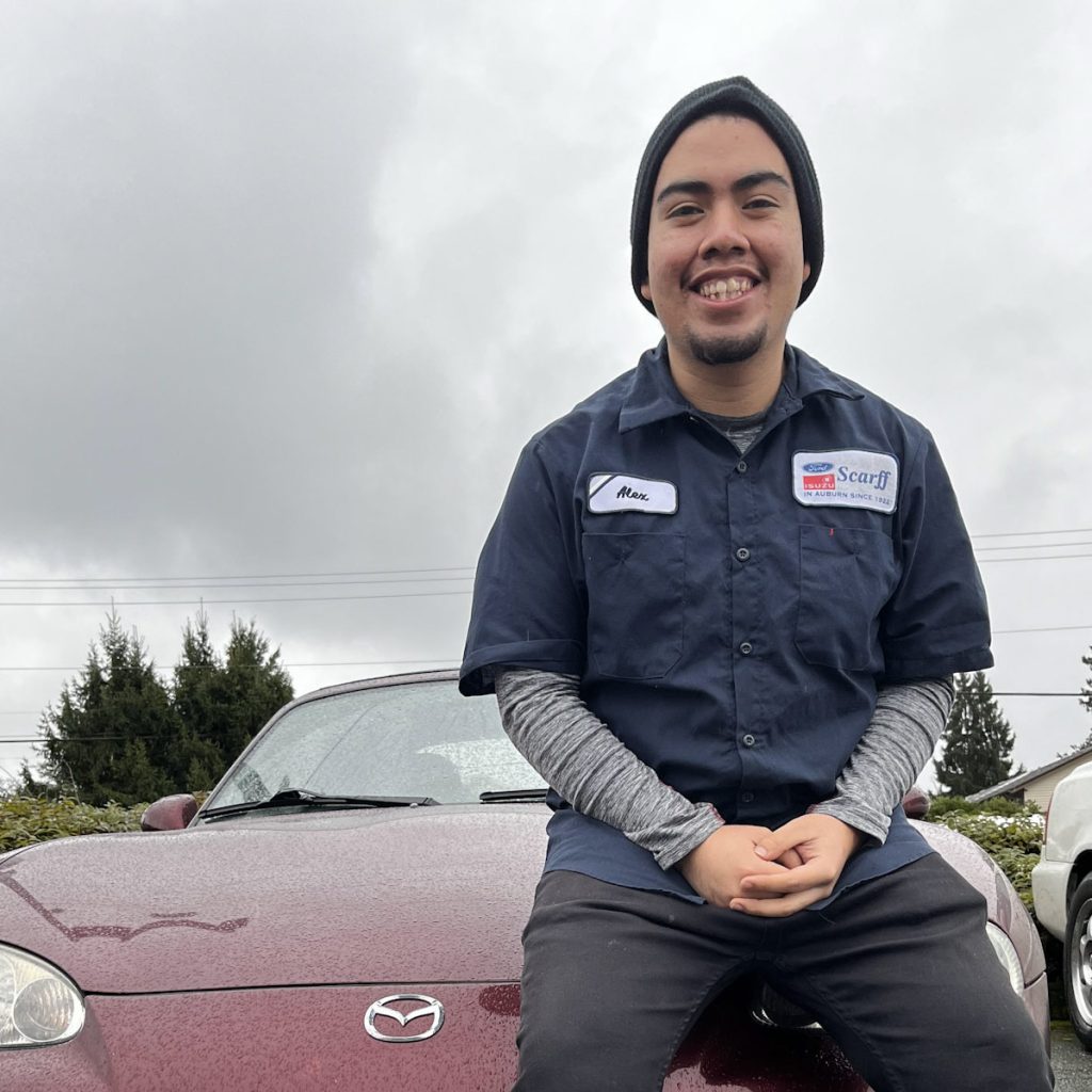 A photo of scholarship recipient Alejandro. Alejandro is wearing a blue technician work shirt, and is sitting on the hood of a red Mazda coupe.