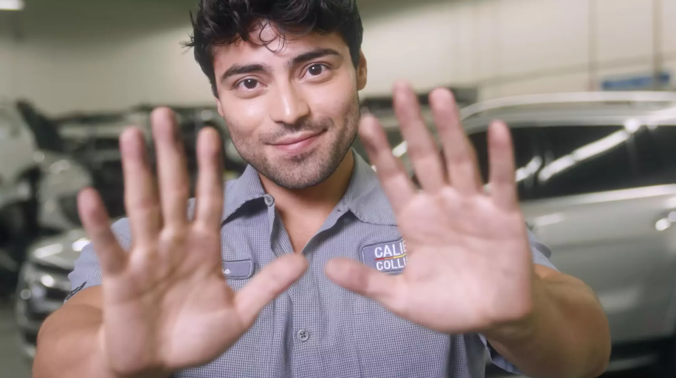 A photo of a collision technician standing in a shop, holding his hands up to the camera and out in front of him while smiling.