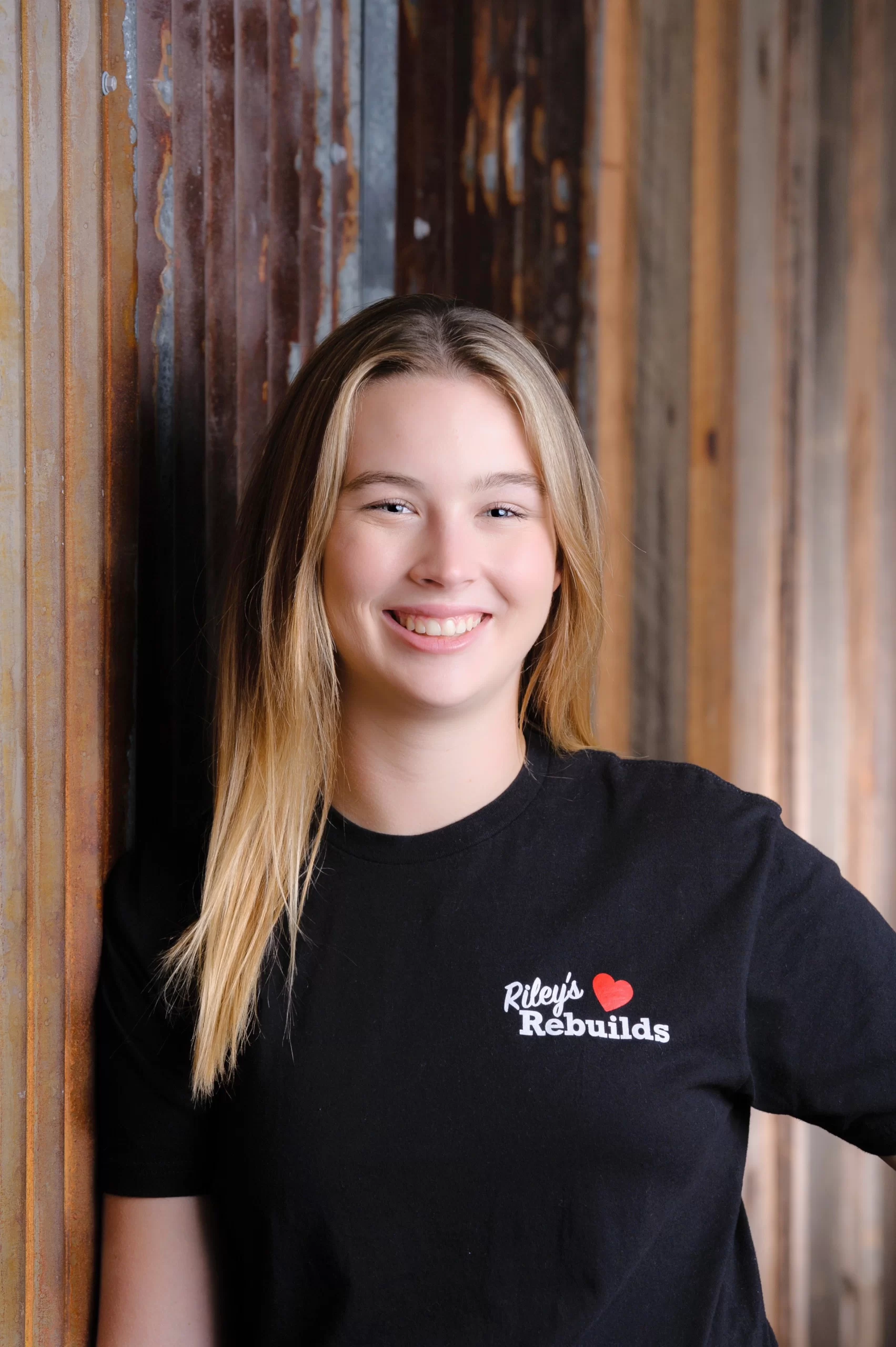 A photo of Riley Schlick-Trask leaning against a wall and smiling. She is wearing a Riley's Rebuilds branded t-shirt.