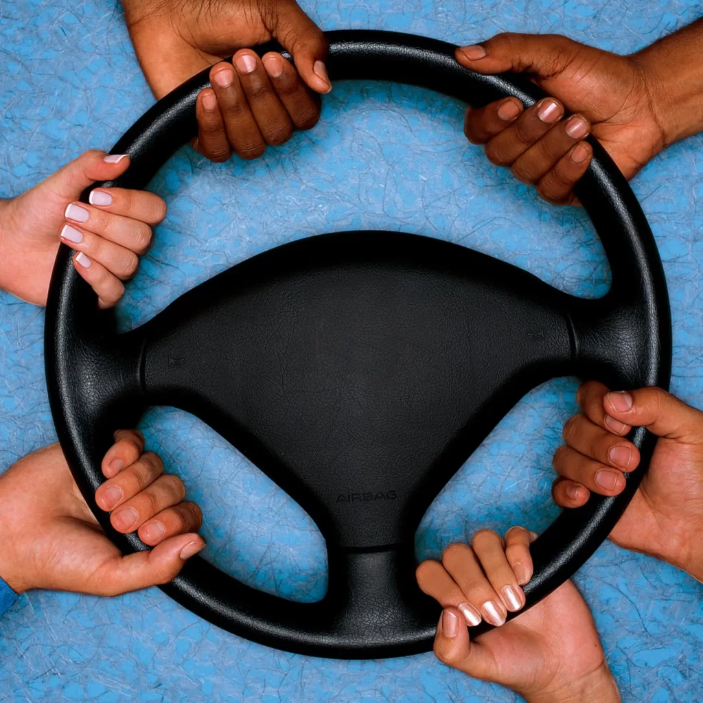 Six hands grasping a steering wheel. The steering wheel is disconnected from any vehicle, and held by the hands over a light blue surface.